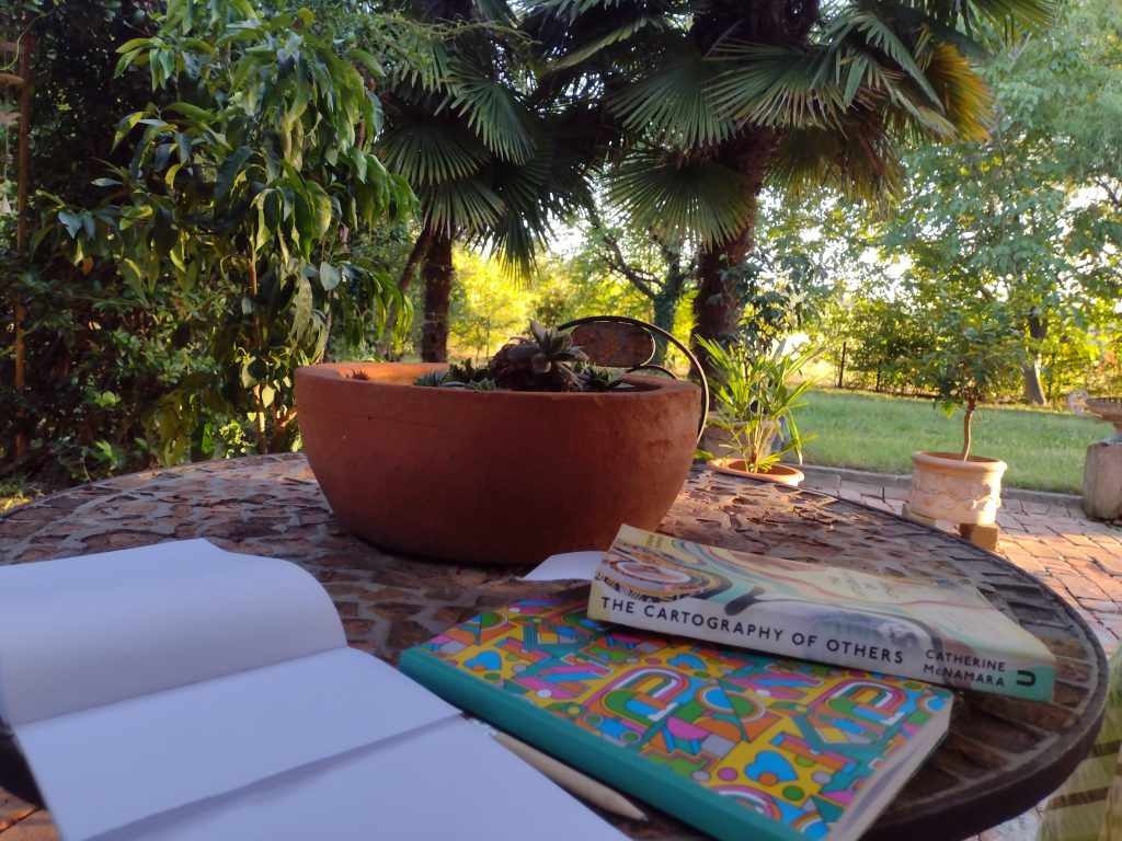 Living with a Writer. In the writer's garden. A table in the sunshine with notebooks and a copy of The Cartography of Others by Catherine McNamara